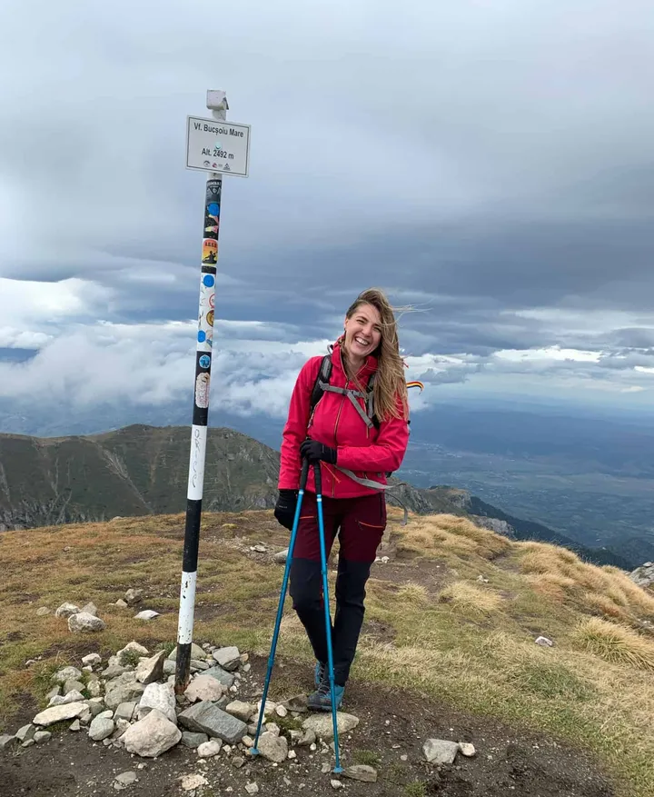 Andreea Giurgilă zâmbind pe vârful unui munte, lângă stâlpul de marcaj turistic, cu bețele de trekking