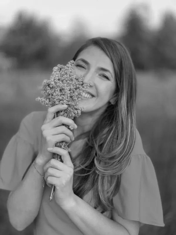 Fotografie alb-negru: Andreea Giurgilă zâmbind pe câmp, cu un buchet de flori lângă obraz