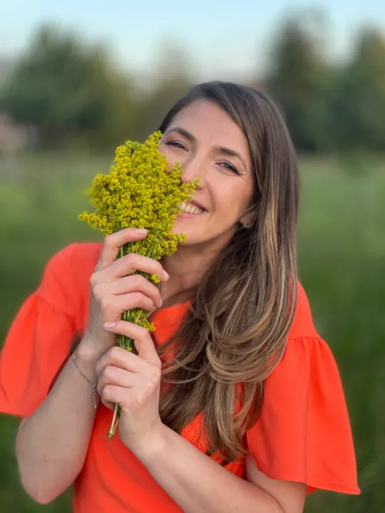 Andreea Giurgilă zâmbind, cu un buchet de flori galbene de câmp în mână, într-o poiană, în bluză portocalie