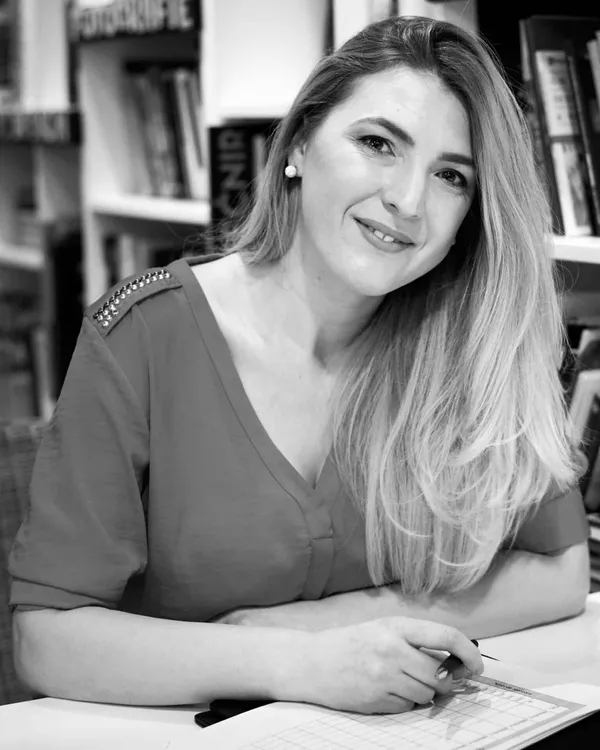 Andreea Giurgilă, life coach acreditat NLP, la birou, zâmbind, cu pixul în mână, în fața unui raft cu cărți, fotografie alb-negru
