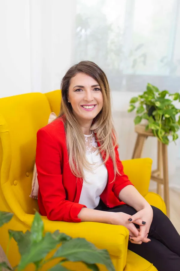 Andreea Giurgilă, life coach în București, în sacou roșu, zâmbind, pe fotoliul galben din cabinet